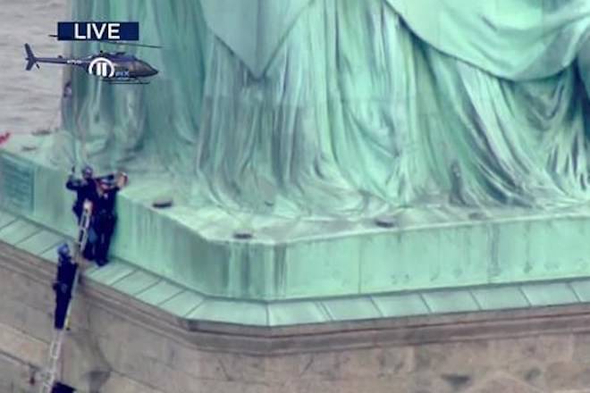 In this image made from television news video by PIX11, a person, right, lies under the right foot of New York’s Statue of Liberty as police officers, left, work to convince the climber to descend, Wednesday, July 4, 2018. Shortly before the climber scaled the statue’s base, forcing its evacuation on the Fourth of July, several people who hung a banner from the Statue of Liberty’s pedestal calling for abolishing the federal government’s chief immigration enforcement agency were arrested. (PIX11 via AP)