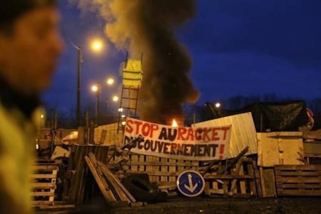 A demonstrator stands in front of a makeshift barricade set up by the so-called yellow jackets to block the entrance of a fuel depot in Le Mans, western France, Tuesday, Dec. 5, 2018, with banner reading “Stop the Government racket”. (AP Photo/David Vincent)